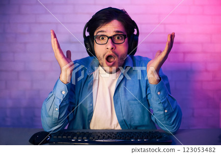 This image features a young man, wearing headphones, sitting in front of a computer and looking shocked and surprised. He is wearing a blue shirt and appears to be playing a video game. This image features a young man, wearing headphones, sitting in front of a computer and looking shocked and surprised. He is wearing a blue shirt and appears to be playing a video game. 123053482