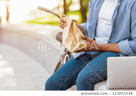 Paper package with lunch in hands of unrecognizable african american man sitting with laptop in park Paper package with lunch in hands of unrecognizable african american man sitting with laptop in park 123053546