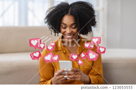 A young black woman with curly hair is sitting at home and smiling while checking her smartphone. She is surrounded by pink heart icons, indicating that she is receiving many likes A young black woman with curly hair is sitting at home and smiling while checking her smartphone. She is surrounded by pink heart icons, indicating that she is receiving many likes 123053555