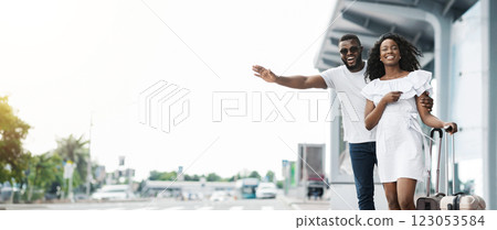 A happy African American couple with a suitcase stands outside an airport terminal, waving their hands at a passing taxi. Couple appears to be ready to start their vacation, copy space A happy African American couple with a suitcase stands outside an airport terminal, waving their hands at a passing taxi. Couple appears to be ready to start their vacation, copy space 123053584