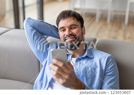 Smiling millennial bearded man reclining on couch at home, using smartphone, scrolling or chatting online 123053607