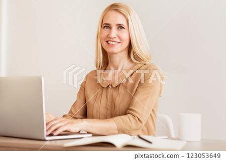 A woman with blonde hair smiles while working on a laptop at a desk, with a notebook and pen nearby. 123053649