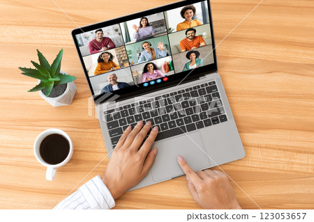A person is sitting at a wooden desk, using a laptop to participate in a video conference. There are multiple participants visible on the screen, top view A person is sitting at a wooden desk, using a laptop to participate in a video conference. There are multiple participants visible on the screen, top view 123053657