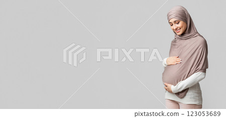 Enjoying pregnancy. Pregnant muslim woman in headscarf tenderly touching her belly, embracing tummy over gray background with empty space, panorama 123053689