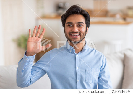 Arab man in a light blue shirt smiles and waves hello with his right hand, standing indoors in a home setting with a white couch partially visible in the background. Arab man in a light blue shirt smiles and waves hello with his right hand, standing indoors in a home setting with a white couch partially visible in the background. 123053699