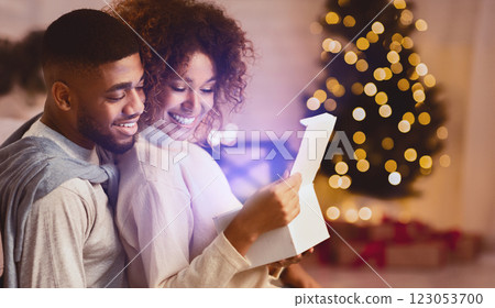 Christmas gift with love. Newlyweds opening gift box together, sitting against xmas tree Christmas gift with love. Newlyweds opening gift box together, sitting against xmas tree 123053700