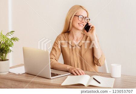 A woman wearing glasses sits at a desk with a laptop, notebook, and pen, talking on a phone while smiling. 123053707