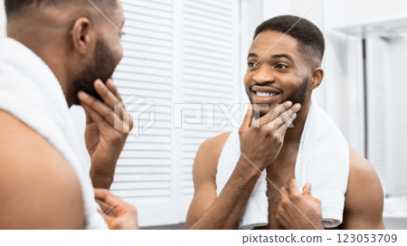 Happy afro man with bare torso touching beard looking in mirror at bathroom. Modern man in bathroom concept 123053709