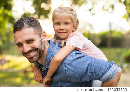 A father and his young daughter are enjoying a day at the park. The father is giving his daughter a piggyback ride, and both of them are smiling and laughing. 123053733