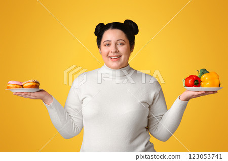 Smiling Plus Sized Girl Holding Plates With Fresh Vegetables And Donuts In Both Hands, Posing Over Yellow Background, Panorama Smiling Plus Sized Girl Holding Plates With Fresh Vegetables And Donuts In Both Hands, Posing Over Yellow Background, Panorama 123053741