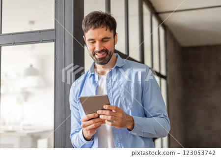 A man in a blue shirt stands in an office building, using a tablet device while looking at the screen with a smile. The man is in a casual work environment, surrounded by large windows. A man in a blue shirt stands in an office building, using a tablet device while looking at the screen with a smile. The man is in a casual work environment, surrounded by large windows. 123053742