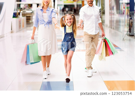 A family of three, a young girl with pigtails and her parents, are walking together through a modern shopping mall. The little girl holds onto both her mom and dad's hands A family of three, a young girl with pigtails and her parents, are walking together through a modern shopping mall. The little girl holds onto both her mom and dad's hands 123053754