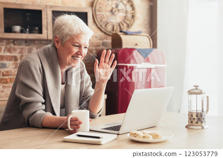 A senior woman sits at a kitchen table with a laptop in front of her. She is holding a cup of coffee in her left hand and waving with her right hand, indicating she is video chatting with someone. 123053779