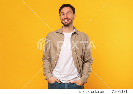 A man wearing a brown button-up shirt and jeans stands against a yellow background. He is smiling directly at the camera with his hands in his pockets. A man wearing a brown button-up shirt and jeans stands against a yellow background. He is smiling directly at the camera with his hands in his pockets. 123053811