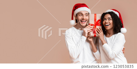 Portrait of happy multiracial couple holding gift box and wearing santa hats, excited what's inside of present Portrait of happy multiracial couple holding gift box and wearing santa hats, excited what's inside of present 123053835