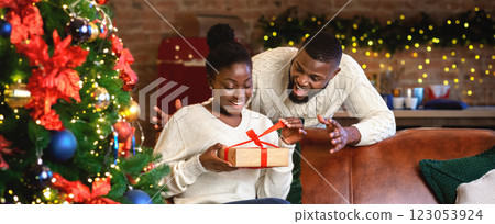 Excited afro wife unwrapping christmas present box from her husband, enjoying holidays together in decorated living room. 123053924