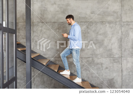 A man in a light blue shirt and jeans walks up a modern concrete staircase while looking at his smartphone. 123054040