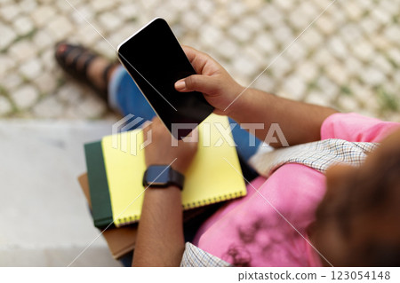 A person is seated on a set of steps, focused on their smartphone while jotting down notes in a yellow notebook. It appears to be a casual outdoor setting, emphasizing productivity. A person is seated on a set of steps, focused on their smartphone while jotting down notes in a yellow notebook. It appears to be a casual outdoor setting, emphasizing productivity. 123054148