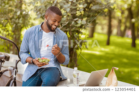 Lunch Fun. Young Black Guy Eating Takeaway Salad Outdoors And Watching Videos On Laptop, Relaxing On Bench In Park 123054401