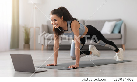 Cheerful active woman doing fitness at home, using laptop, watching online lessons. Positive well-fit lady in sportswear exercising during covid-19 pandemic, watching fitness videos on Internet 123054402
