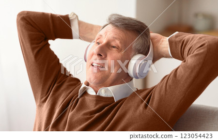 Relaxed Senior Man In Headphones Listening To Music Holding Hands Behind Head Sitting On Sofa At Home. Selective Focus Relaxed Senior Man In Headphones Listening To Music Holding Hands Behind Head Sitting On Sofa At Home. Selective Focus 123054423