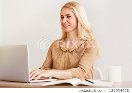A blonde woman sits at a desk with a laptop, smiling as she types. 123054458