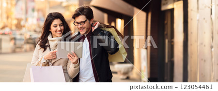 A couple with shopping bags smiles while looking at a tablet outdoors on a busy city street, copy space 123054461