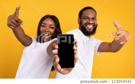 Advertising New Mobile App. Portrait of happy black couple holding smartphone with empty screen in hands, showing and pointing at device with blank space for mockup, yellow studio background Advertising New Mobile App. Portrait of happy black couple holding smartphone with empty screen in hands, showing and pointing at device with blank space for mockup, yellow studio background 123054541