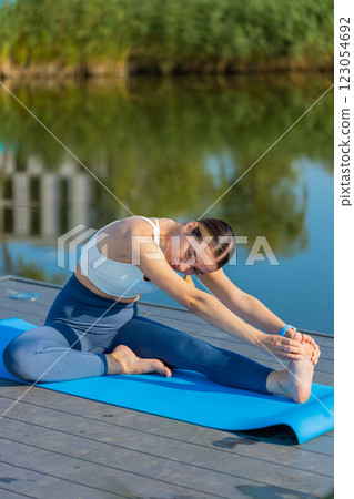 Fit Caucasian young woman in sportswear doing stretching exercising on yoga mat near pond in park 123054692