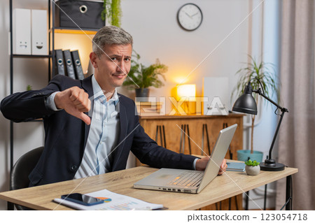 Upset young Caucasian freelancer in suit using laptop and showing thumb down at home office table Upset young Caucasian freelancer in suit using laptop and showing thumb down at home office table 123054718