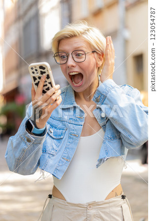Happy Caucasian blonde woman using smartphone celebrating win good message news on city town street 123054787