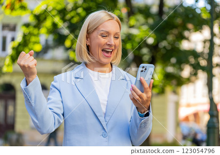 Mature Caucasian businesswoman CEO celebrating good news on smartphone standing on city street Mature Caucasian businesswoman CEO celebrating good news on smartphone standing on city street 123054796