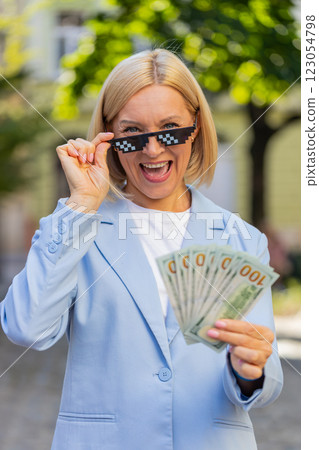 Happy rich mature business woman putting on thug life sunglasses showing dollar money on city street 123054798