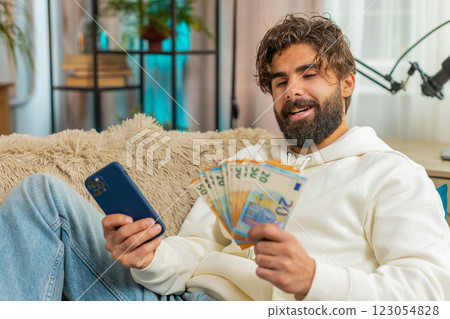 Successful rich young Indian man holding euro bill and smartphone sitting on sofa couch at home 123054828