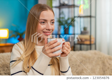 Caucasian woman enjoying hot tea on sofa holding cup both hands relaxing indoors calm comfortable Caucasian woman enjoying hot tea on sofa holding cup both hands relaxing indoors calm comfortable 123054876