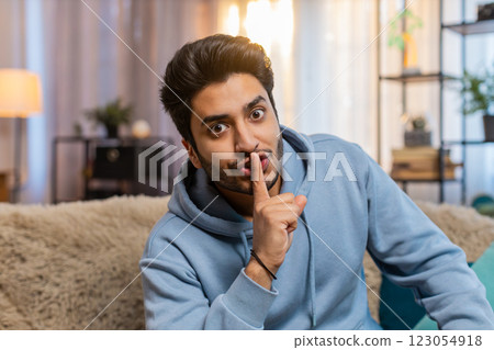 Young Indian man pressing index finger to lips making silence gesture sitting on sofa couch at home 123054918