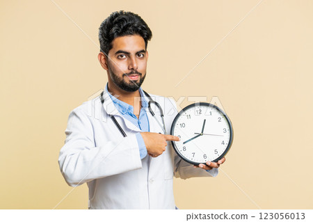 Doctor man showing time on wall office clock, ok, thumb up, approve, pointing finger at camera Doctor man showing time on wall office clock, ok, thumb up, approve, pointing finger at camera 123056013