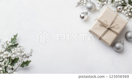 A wrapped gift with twine lies on a white surface, surrounded by silver ornaments and sprigs of greenery with white berries. A wrapped gift with twine lies on a white surface, surrounded by silver ornaments and sprigs of greenery with white berries. 123056569