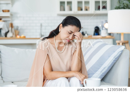 Upset Asian senior woman sitting on sofa, lost in thoughts, feeling lonely 123058636
