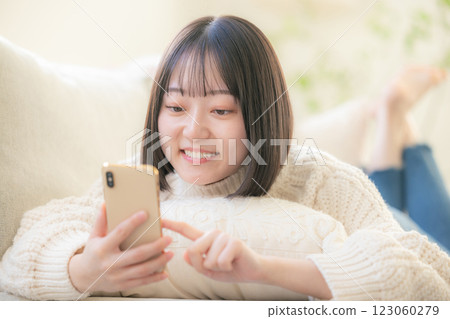 一位年輕女子躺在沙發上，看著她的智慧型手機 123060279
