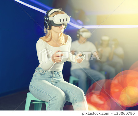In darkened hall of gaming club, girl in VR helmet takes part in group game 123060790