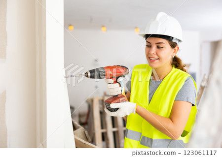 Woman builder in a protective helmet and a yellow vest drills hole with a screwdriver Woman builder in a protective helmet and a yellow vest drills hole with a screwdriver 123061117