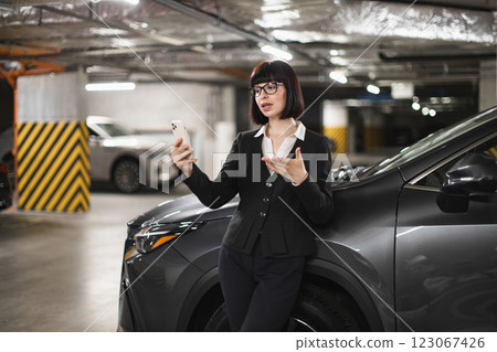 Young adult businesswoman using smartphone for online video call in indoor car parking. Confident professional wearing formal attire, standing beside luxury vehicle. Technology and communication theme 123067426