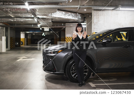 Confident woman standing by car in parking garage, holding phone, conducting online video call. Professional attire, smiling expression, modern workplace scenario. 123067430