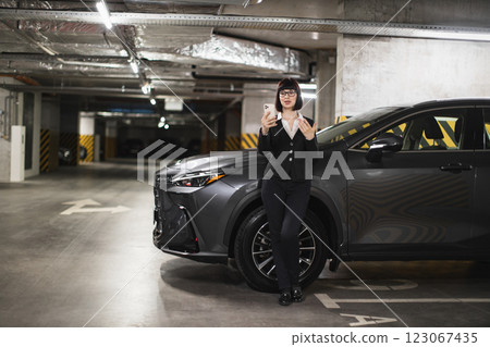 Confident Caucasian businesswoman using smartphone for video call in underground car park. Middle-aged woman in office attire near modern vehicle, engaging in online communication. Confident Caucasian businesswoman using smartphone for video call in underground car park. Middle-aged woman in office attire near modern vehicle, engaging in online communication. 123067435