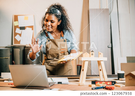 Carpenter woman in a workshop holding furniture design sketches, presenting her project to a customer using a laptop, surrounded by unfinished wooden pieces and tools, craftsmanship and creativity 123067938