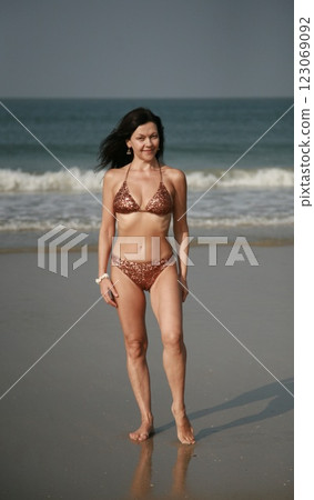 Pretty woman 51 years old in a fashionable swimsuit with sequins stands on the beach against the backdrop of ocean waves Goa India Pretty woman 51 years old in a fashionable swimsuit with sequins stands on the beach against the backdrop of ocean waves Goa India 123069092