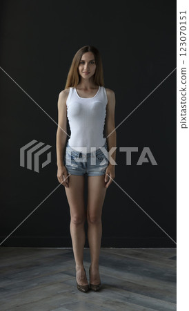 young woman in a white tank top and denim shorts on a dark background, full length portrait 123070151