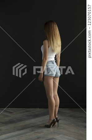 young woman in a white tank top and denim shorts on a dark background, full length portrait 123070157
