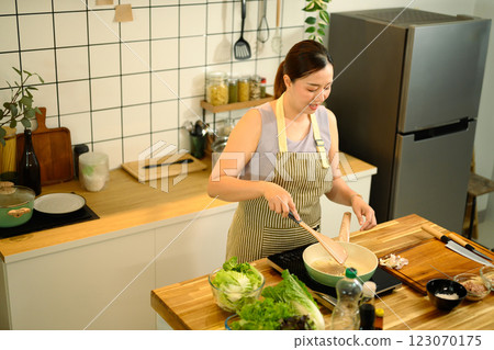 Smiling middle aged woman stirring ingredients in frying pan while preparing meal in cozy kitchen Smiling middle aged woman stirring ingredients in frying pan while preparing meal in cozy kitchen 123070175